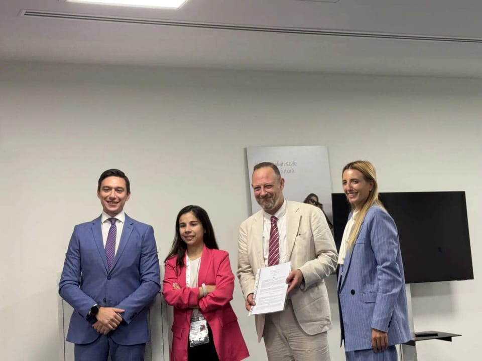 Quattro persone in piedi in un ufficio: due uomini in completo, una donna con giacca rosa e una donna in completo azzurro; l’uomo con la giacca beige tiene in mano un foglio.
