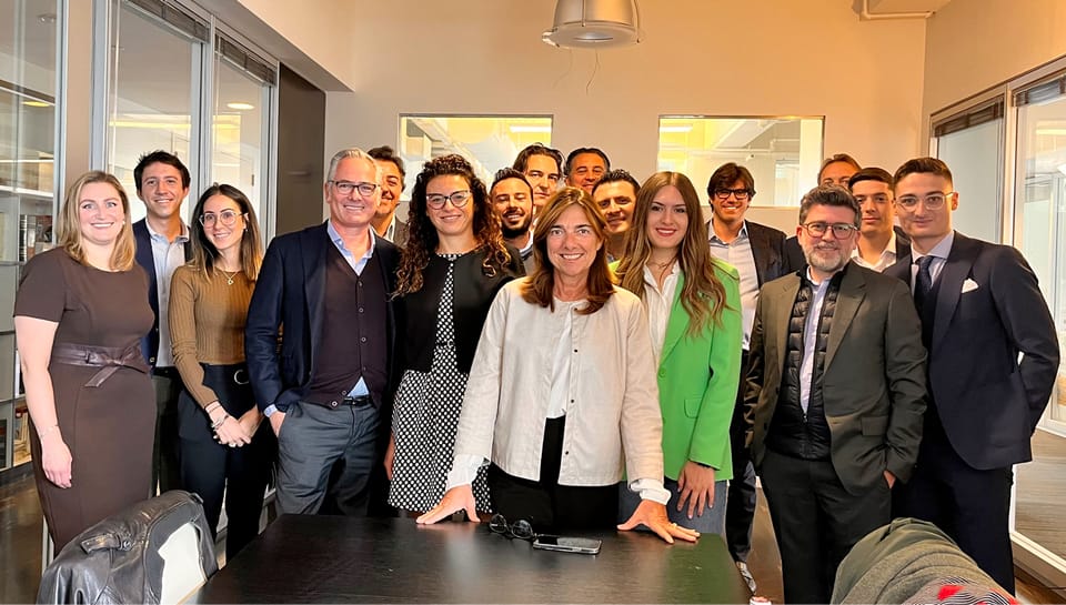 Gruppo numeroso di persone sorridenti in posa in un ufficio con grandi vetrate; al centro una donna con camicia bianca appoggia le mani su un tavolo scuro.