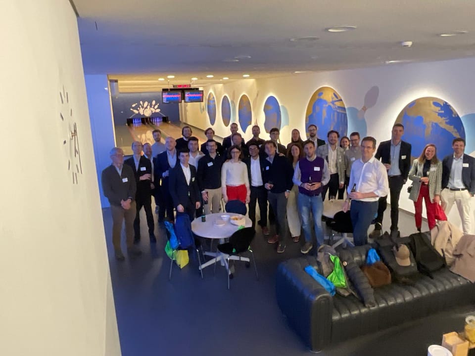 Un grande gruppo di persone in piedi in una sala dalle pareti blu con grandi disegni di mappamondi; in primo piano un tavolino rotondo e dei divani con delle borse.