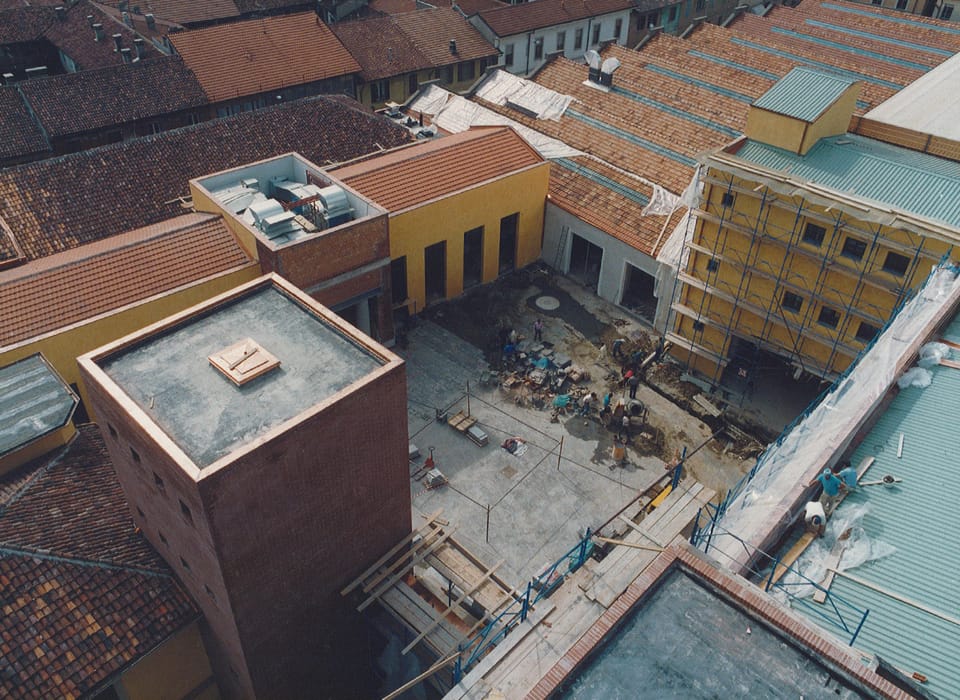 Veduta dall’alto di un cantiere tra tetti di tegole: in primo piano una torre di mattoni a pianta quadrata, a destra un edificio giallo coperto da ponteggi, al centro un cortile con operai e materiali.