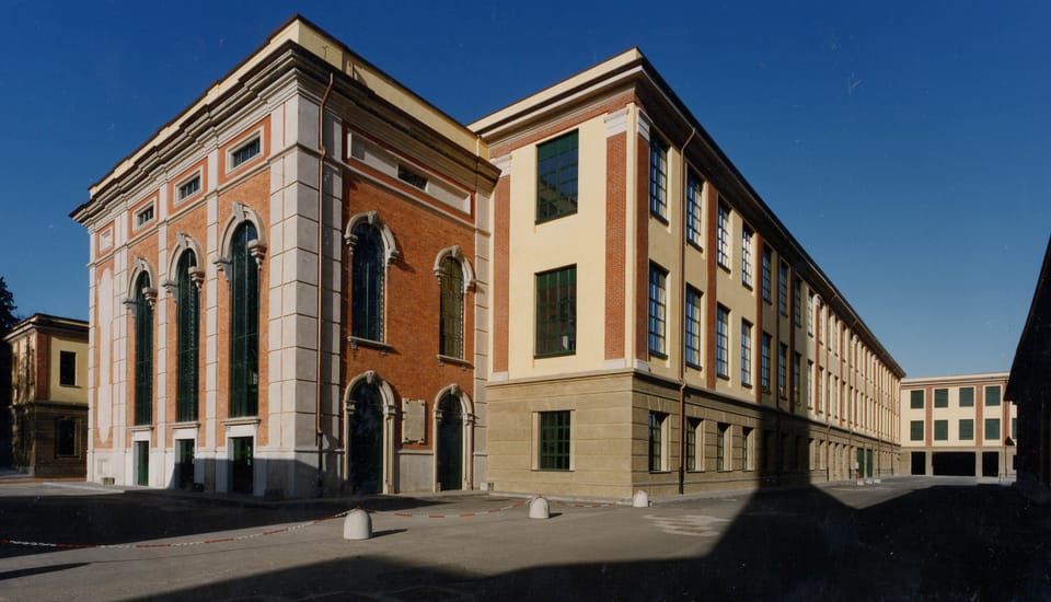 Un edificio storico con facciata in mattoni rossi, lesene chiare e alte finestre ad arco, affiancato da un lungo corpo di fabbrica a tre piani con intonaco chiaro; cielo blu intenso e piazzale in ombra in primo piano.
