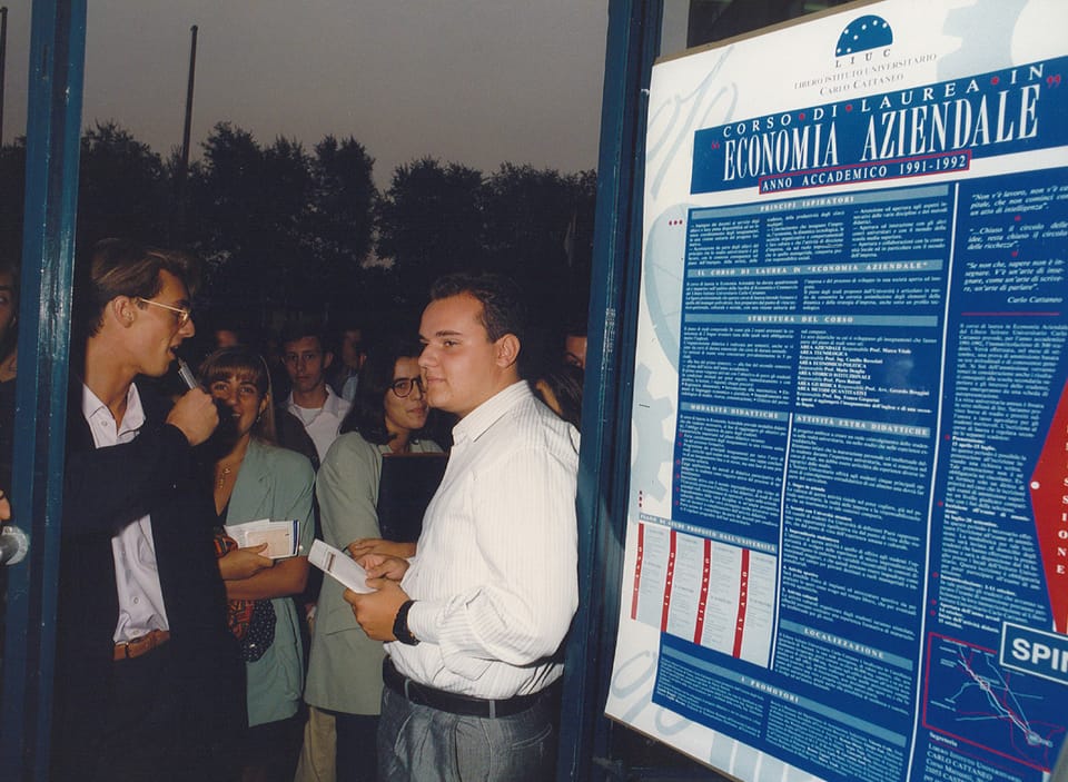 Un giovane con occhiali tiene un microfono davanti a un ragazzo in camicia bianca; attorno altri giovani. A destra un grande manifesto blu: «Corso di laurea in Economia Aziendale, anno accademico 1991-1992».