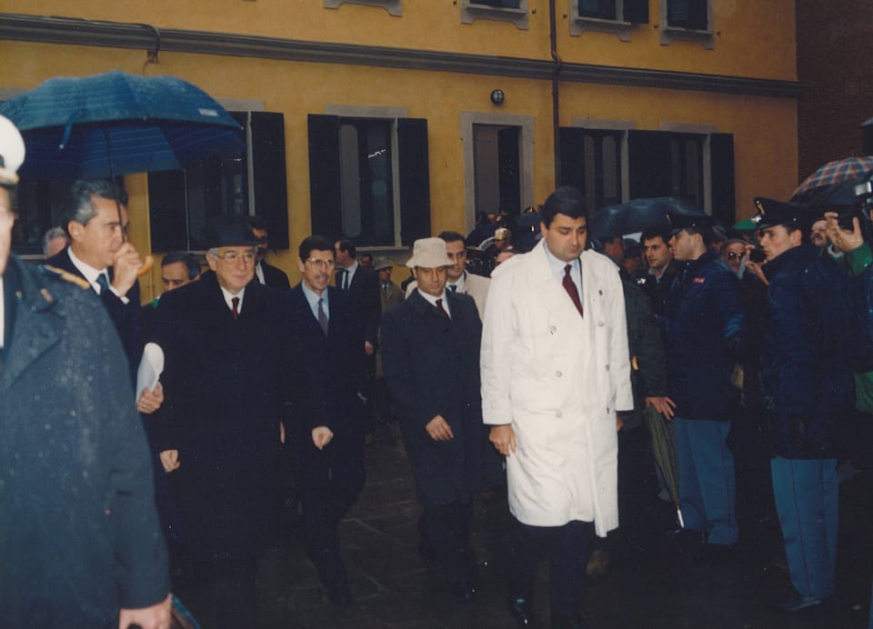 Un gruppo di uomini in cappotto scuro cammina in un cortile bagnato davanti a un edificio giallo; uno indossa un impermeabile bianco, altri reggono ombrelli; ai lati agenti in uniforme e un operatore con la telecamera.