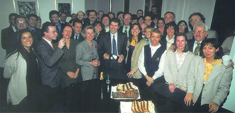 Una trentina di persone, uomini e donne in abiti da ufficio, sorridono raccolte attorno a un tavolo scuro con vassoi di dolci e bicchieri.