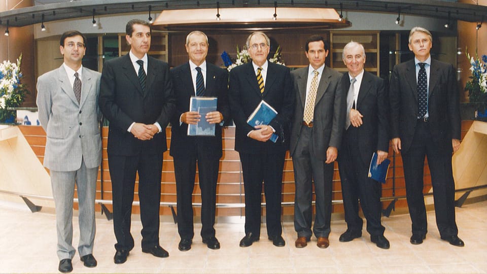 Sette uomini in abito scuro o grigio in posa in fila in un atrio con pavimento chiaro e bancone in legno; tre tengono in mano un volume blu; ai lati composizioni di fiori.