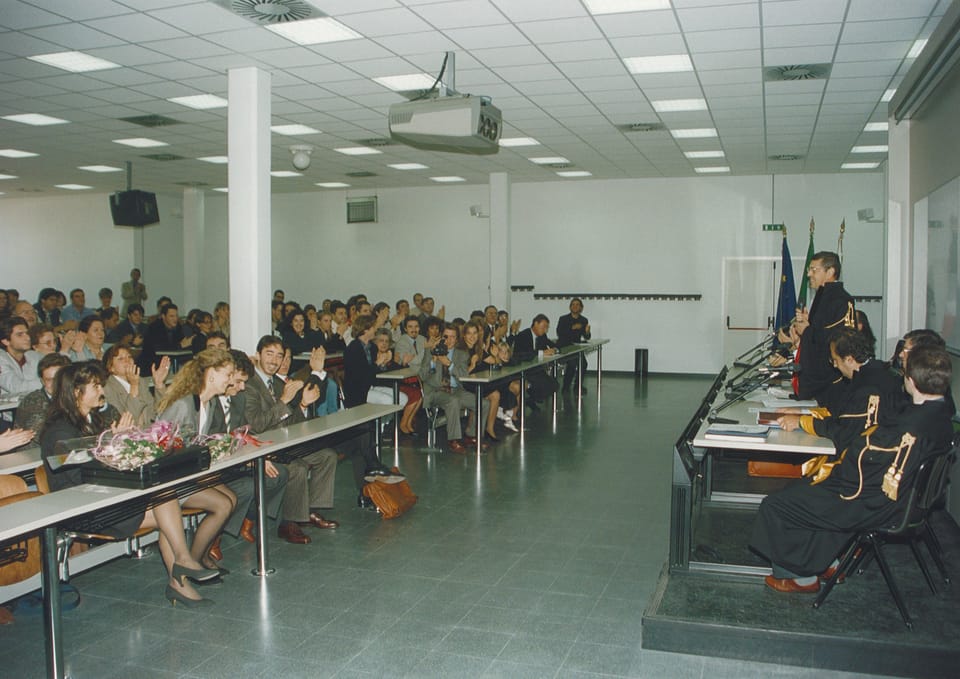 Un’aula luminosa con soffitto a pannelli e un proiettore: a sinistra il pubblico seduto ai banchi applaude, con mazzi di fiori sul tavolo; a destra, su una pedana, la commissione in toga nera e un docente in piedi al microfono.