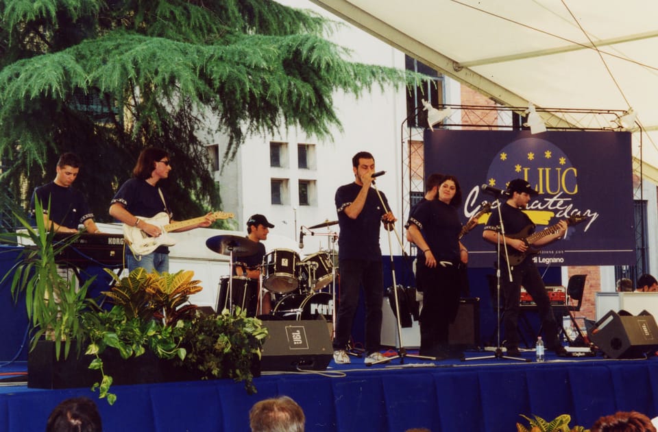 Sei musicisti in maglietta scura su un palco con tastiera, chitarre, batteria e microfoni; alle spalle un fondale blu con la scritta «LIUC Graduate day»; in primo piano piante e il bordo blu del palco.