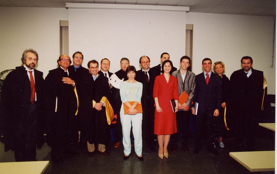 Foto di gruppo in un’aula: una decina di docenti in toga nera con mantelline gialle e alcuni giovani in abito, tra cui una donna vestita di rosso e una in abito chiaro, con cartelline in mano.