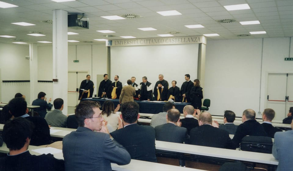Vista dal fondo di un’aula: il pubblico seduto ai banchi applaude; in fondo la commissione in toga nera in piedi dietro un tavolo blu, sotto un’insegna con la scritta «Università Cattaneo Castellanza».