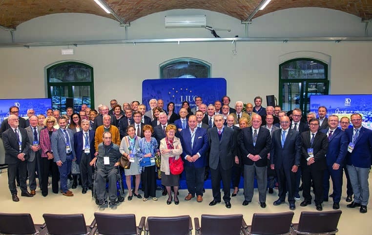 Foto di gruppo di circa settanta persone in una sala con soffitto a volte in mattoni e finestre ad arco; al centro un pannello blu con il logo LIUC, in primo piano una fila di sedie vuote.