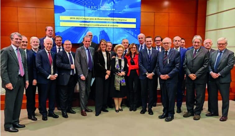 Una trentina di persone in abito scuro in posa davanti a una parete in legno rossiccio; sullo schermo alle loro spalle la scritta «2018-2023 Cinque anni di Osservatorio Banca Impresa».