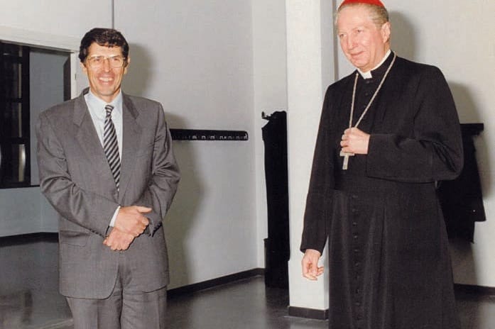 Due uomini in piedi in un corridoio luminoso: a sinistra uno in giacca grigia e cravatta, con le mani incrociate; a destra un ecclesiastico con veste nera, calotta rossa e croce pettorale.