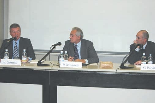 Tre uomini seduti dietro un tavolo da conferenza con microfoni e bottigliette; davanti a loro i cartellini con i nomi, tra cui «P. Mennea».
