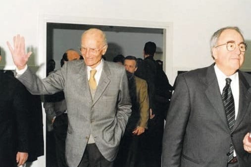 Due uomini anziani in giacca in una sala: uno alza una mano in segno di saluto, l’altro, con occhiali e cravatta, è in primo piano.