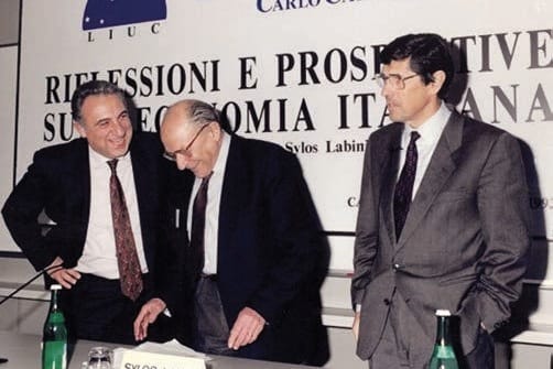 Tre uomini in giacca e cravatta dietro un tavolo con bottiglie d’acqua: due si parlano chinandosi l’uno verso l’altro; alle loro spalle uno striscione con una scritta parziale sull’economia italiana.