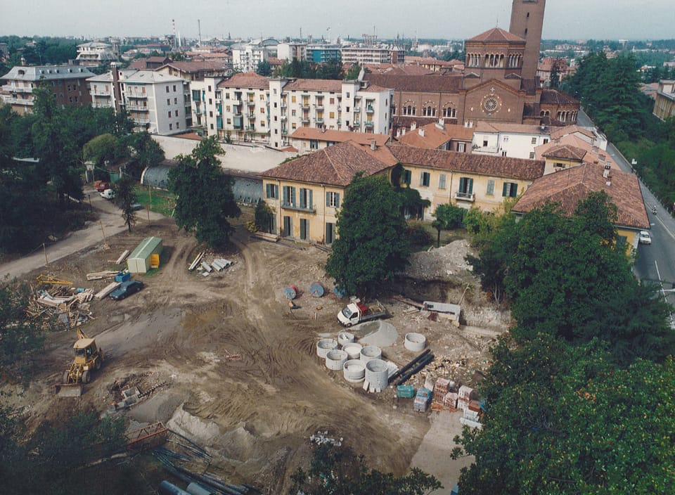Veduta aerea di un cantiere: un’ampia area di terra smossa con anelli di cemento e alcuni mezzi, circondata da alberi; sullo sfondo un edificio giallo storico e, più lontano, le case della città con un campanile.