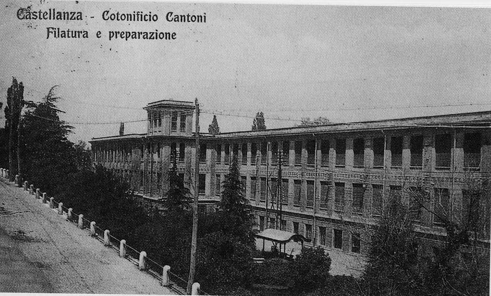 Cartolina d’epoca in bianco e nero con la scritta «Castellanza - Cotonificio Cantoni, Filatura e preparazione»: un lungo edificio di mattoni a più piani con due torrette, un viale con parapetto e alberi in primo piano.