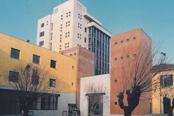 Un cortile tra edifici di mattoni e intonaco giallo e grigio, con una torre bianca a finestre e una torre di mattoni sullo sfondo; in primo piano, a destra, alberi dalle chiome potate; cielo azzurro.