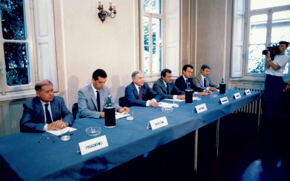 Sala con grandi finestre: sei uomini in giacca siedono a un lungo tavolo con tovaglia blu, ciascuno con un cartellino con il nome (il primo legge «Praderio»); a destra un operatore riprende la scena con la telecamera.