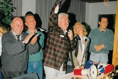 Un gruppetto di persone sorridenti in posa in una sala: un uomo con camicia a quadri alza un braccio, un altro applaude; in primo piano un megafono sul tavolo.