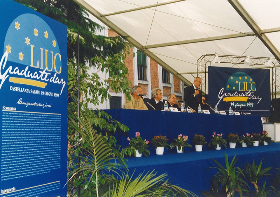 Un palco sotto una tensostruttura bianca con un tavolo coperto da un drappo blu e vasi di fiori; sullo sfondo un edificio di mattoni e striscioni con la scritta «LIUC Graduate day» e la data 19 giugno 1999.