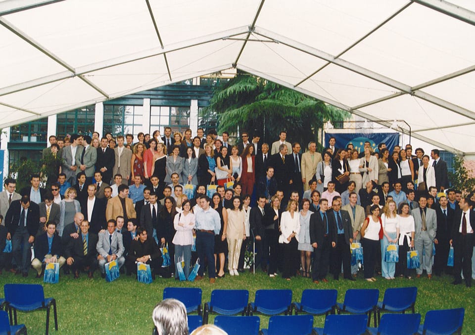 Foto di gruppo sotto una grande tensostruttura bianca: centinaia di laureati e ospiti in piedi sul prato, con sedie blu in primo piano.