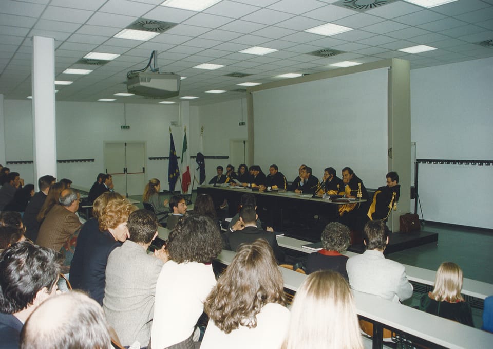 Un’aula con soffitto di pannelli luminosi e un proiettore: in primo piano il pubblico, di spalle, seduto ai banchi; in fondo un lungo tavolo con la commissione in toga e alcune bandiere.