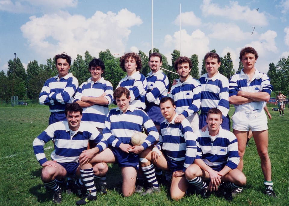 Una squadra di giovani giocatori di rugby in maglia a righe bianche e blu posa in due file sul prato, con alberi sullo sfondo.