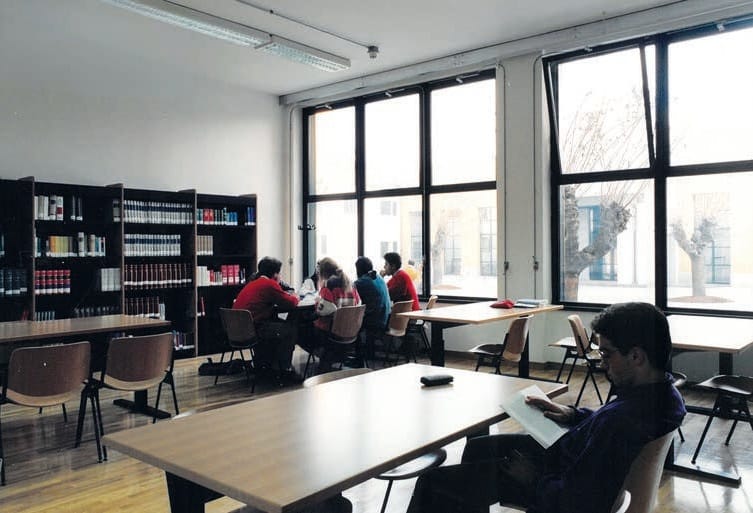 Una sala di biblioteca luminosa con grandi finestre: scaffali di libri a sinistra, tavoli di studio e, in primo piano, un giovane seduto che legge; sullo sfondo altri studenti.