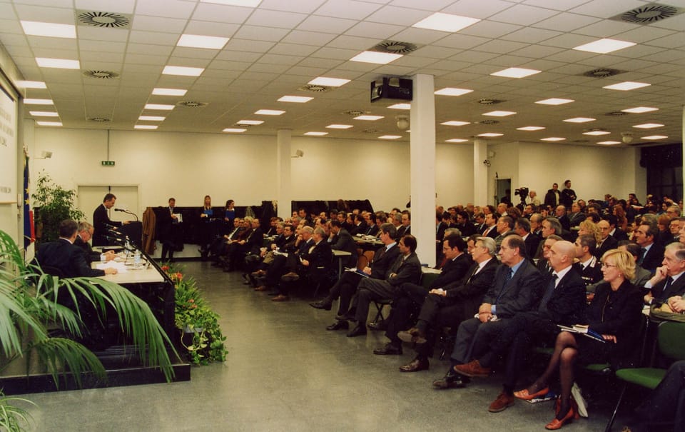 Un’ampia sala conferenze con soffitto a pannelli luminosi: il pubblico, in abiti scuri, siede in file; sul lato sinistro un tavolo di presidenza con un oratore al microfono e piante in primo piano.