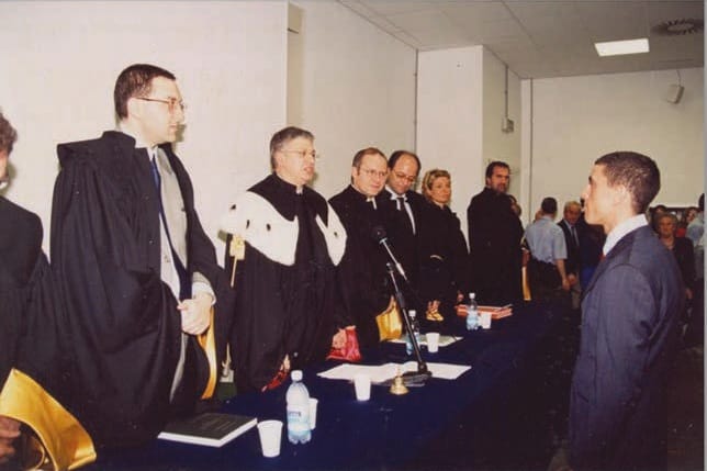 Docenti in toga nera con mantelline bianche in fila dietro un tavolo con tovaglia blu; a destra, davanti a loro, un giovane in abito scuro.