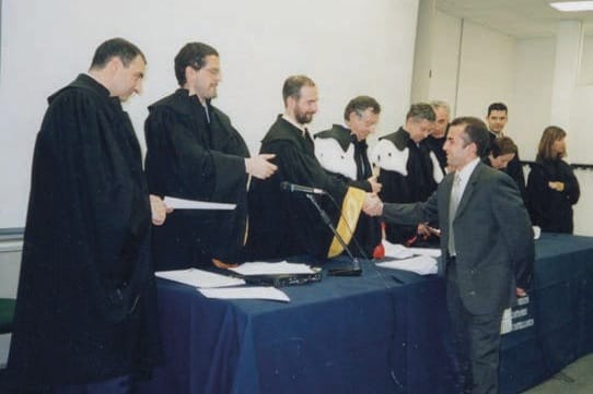Una cerimonia di laurea: alcuni docenti in toga nera dietro un tavolo con tovaglia blu; a destra un uomo in abito grigio stringe la mano a uno di loro.