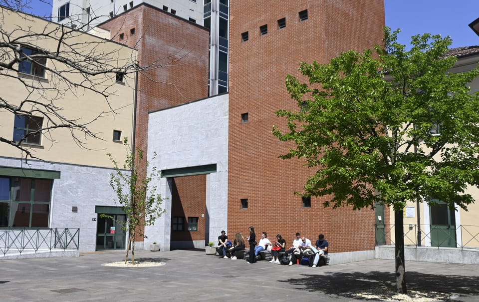 Il cortile del campus in una giornata di sole: un’alta torre di mattoni rossi, edifici con intonaco giallo e grigio, un albero verde a destra e un gruppo di persone sedute su una panchina in fondo.