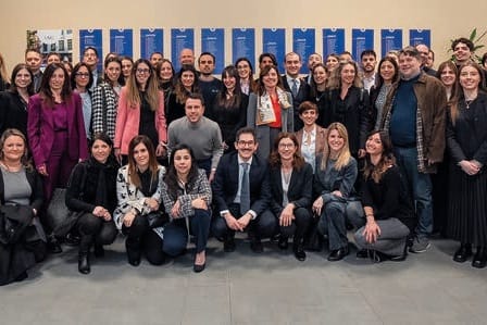Fotografia di gruppo di molte persone, giovani e adulte, in più file davanti a una parete con striscioni blu; in primo piano alcune sono sedute o accovacciate.